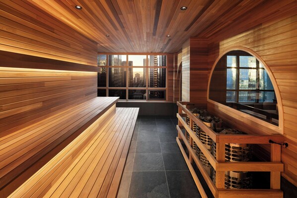 A sauna with wooden walls and benches, a large arched window, and a view of buildings. - The Westin Calgary (Calgary)