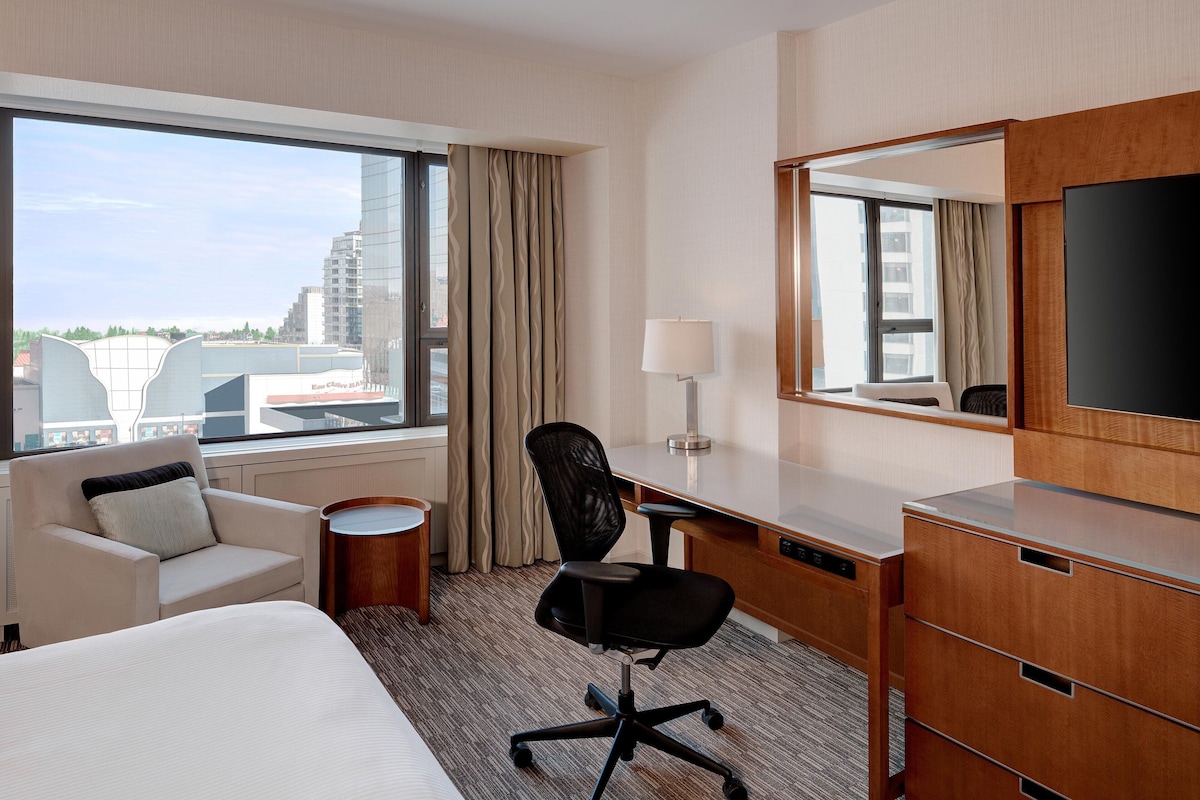 A hotel room with a bed, a desk, a chair, a lamp, a dresser, and a large window with a view of buildings.