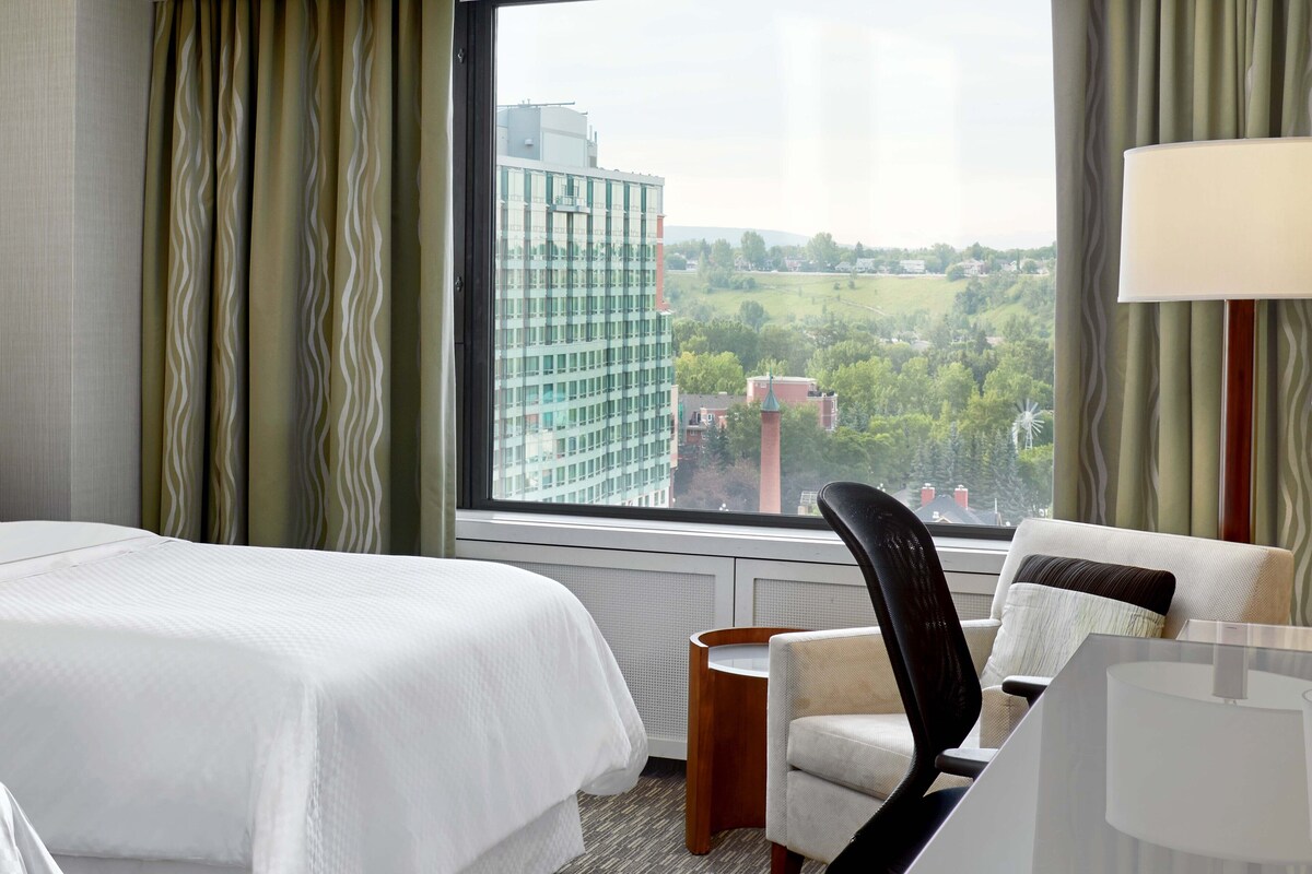 A hotel room with two beds, a chair, a table, and a view of a cityscape through large windows.