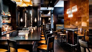 A bar area with a long counter, multiple seating options, and a variety of bottles on shelves. - The Westin Calgary (Calgary)