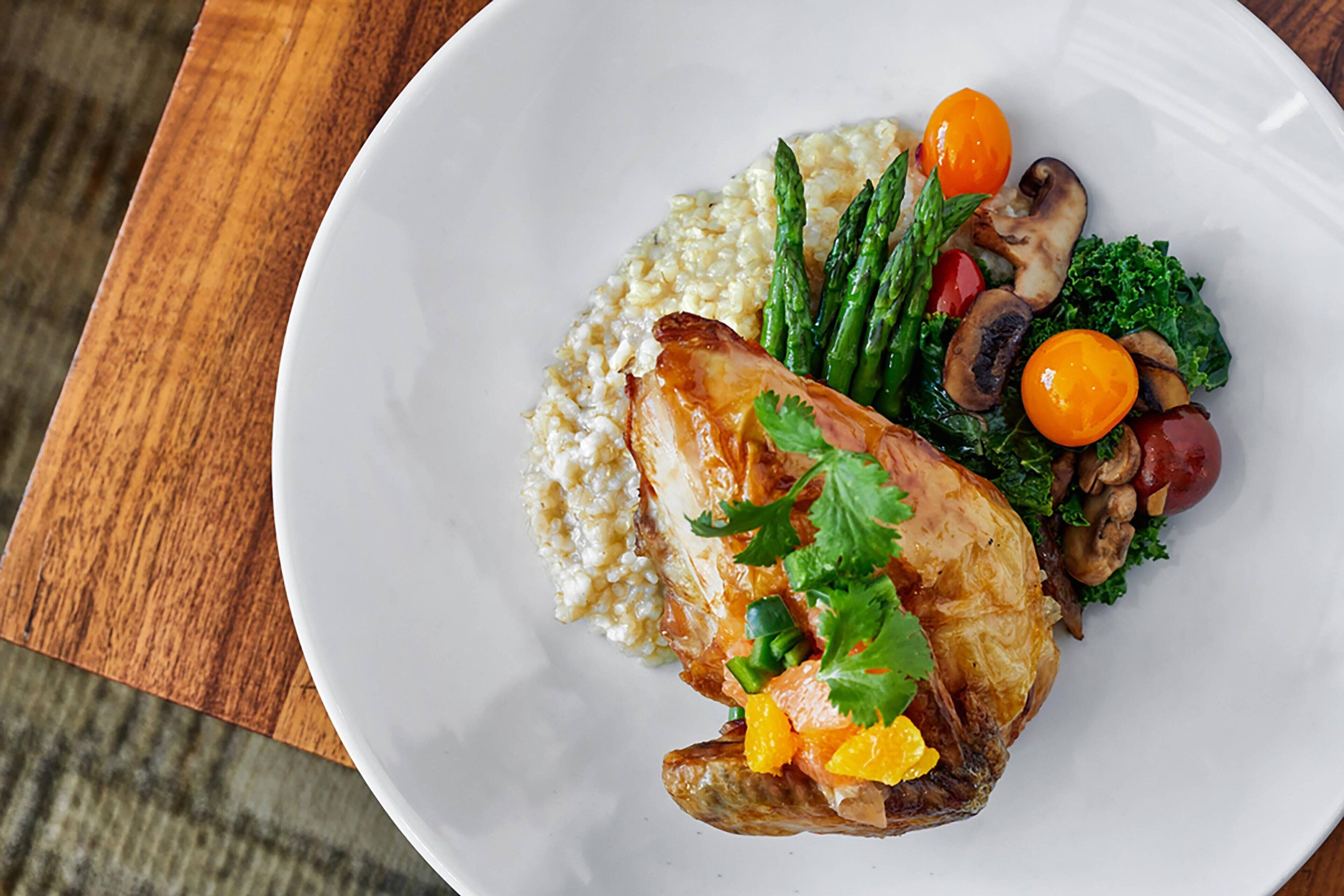 A plate with a roasted chicken, asparagus, cherry tomatoes, mushrooms, kale, and a side of rice.