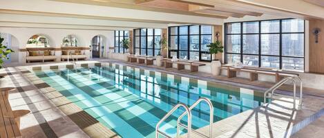 A large indoor swimming pool with clear glass walls, wooden deck, and seating areas with plants.