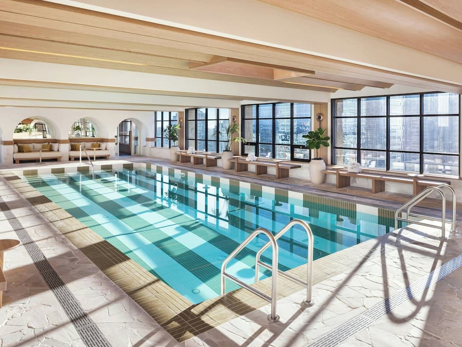 A large indoor swimming pool with clear blue water, surrounded by sunlit seating areas and large windows offering views of the city.