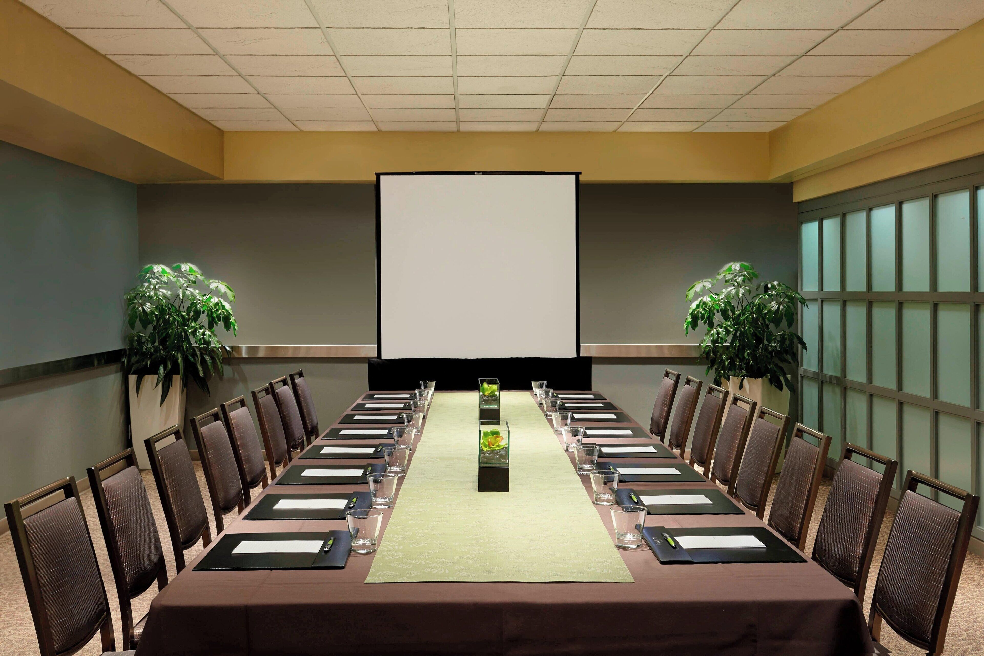 A conference room with a long table, chairs, a projector screen, and potted plants.