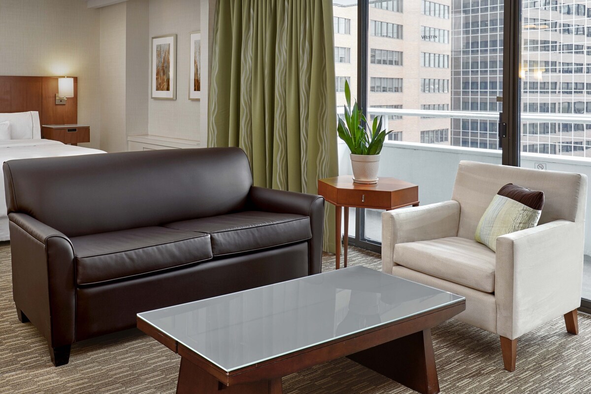 A hotel room with a brown leather sofa, a beige armchair, a small wooden table, and a large window offering a city view.