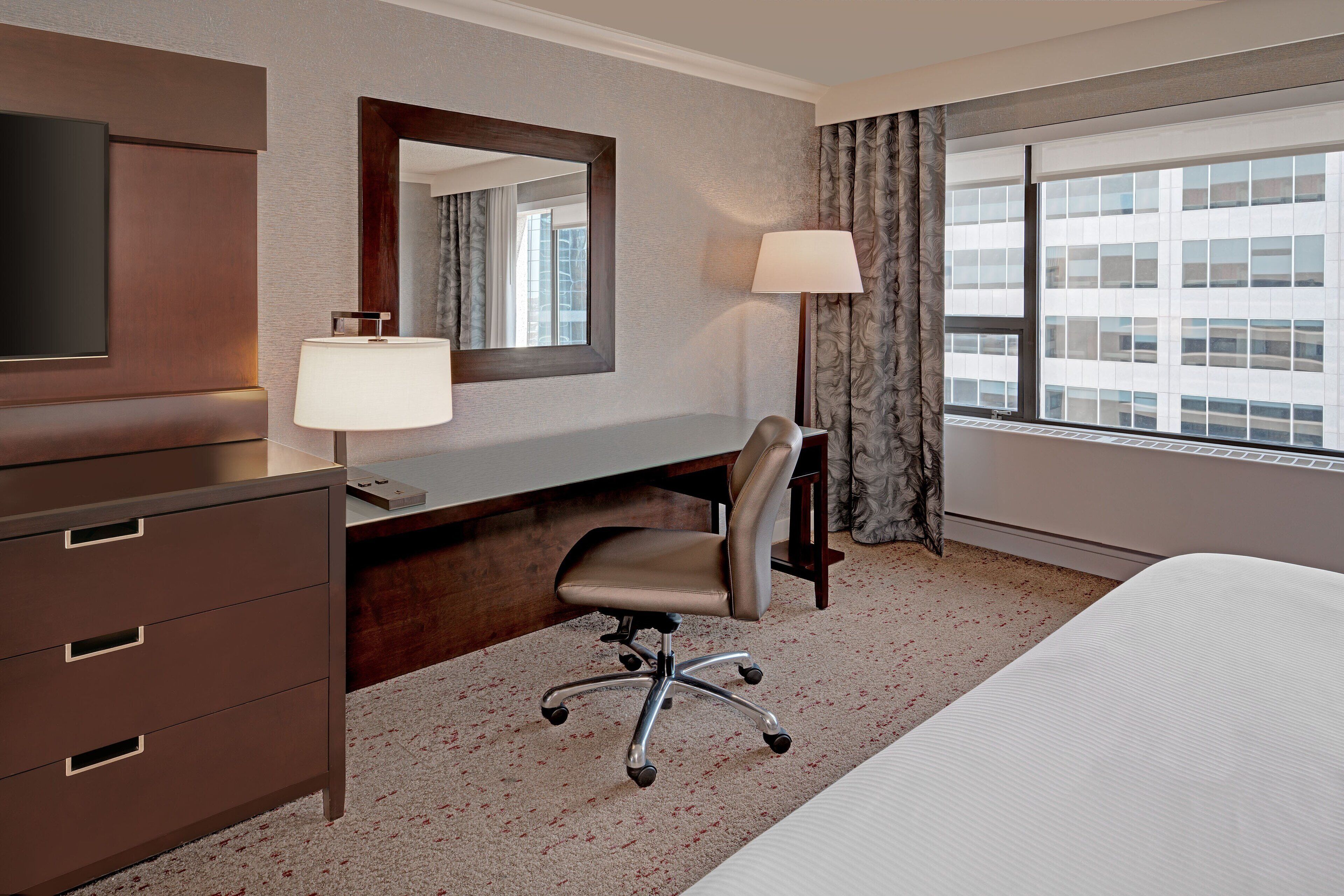 A hotel room with a bed, a desk with a lamp, a chair, a dresser, a large mirror, and a window with curtains.