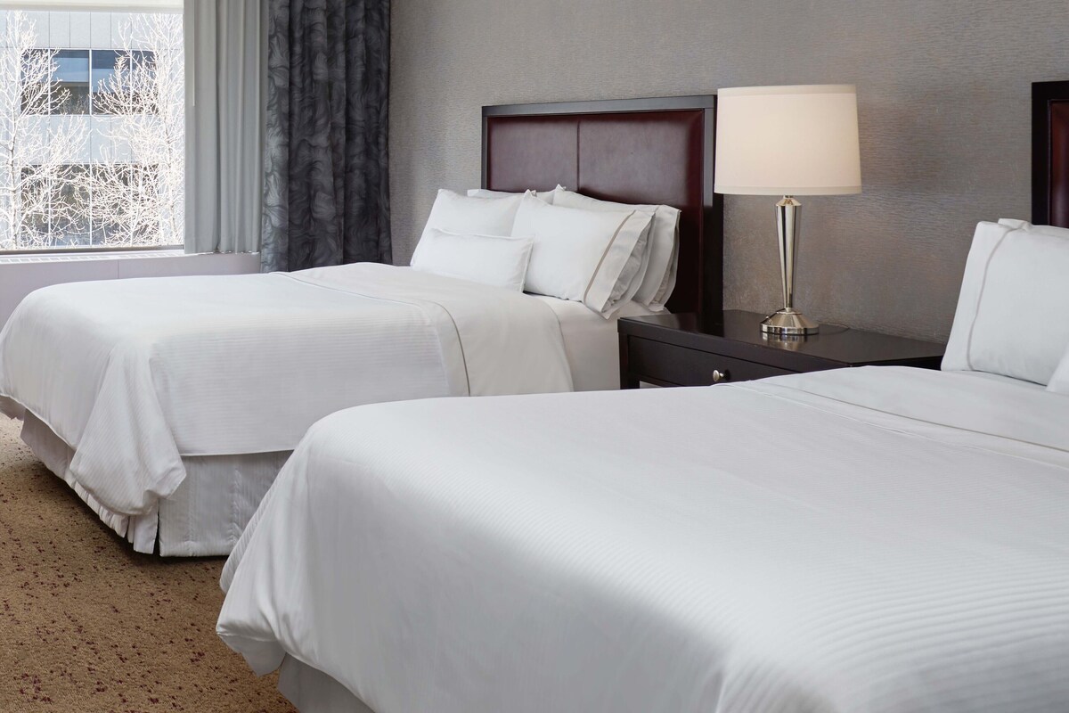 A hotel room with two beds, a nightstand, a lamp, and a window with curtains.