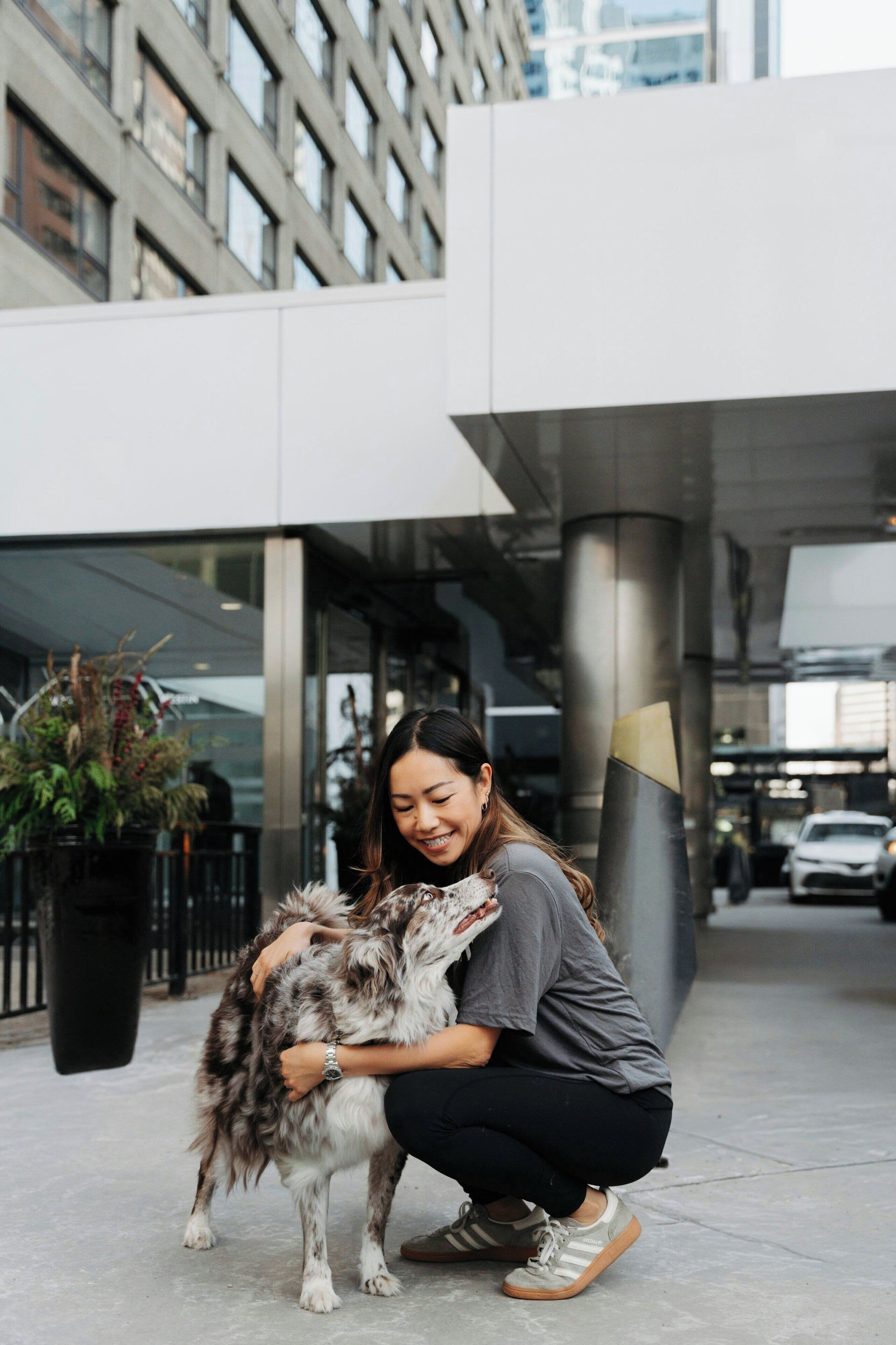 A person crouching next to a dog in an urban setting with modern buildings in the background.