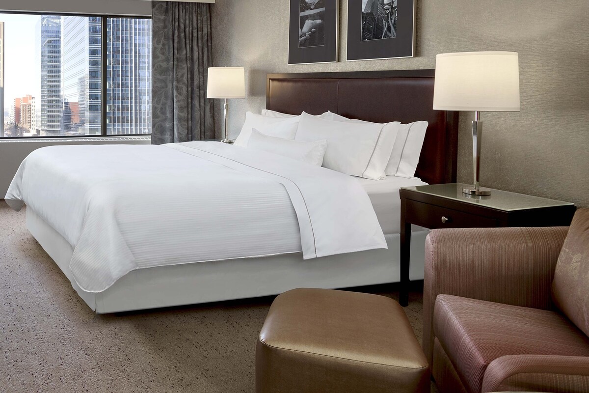 A hotel room with a large bed, two bedside lamps, a brown armchair, and a view of city buildings through the window.