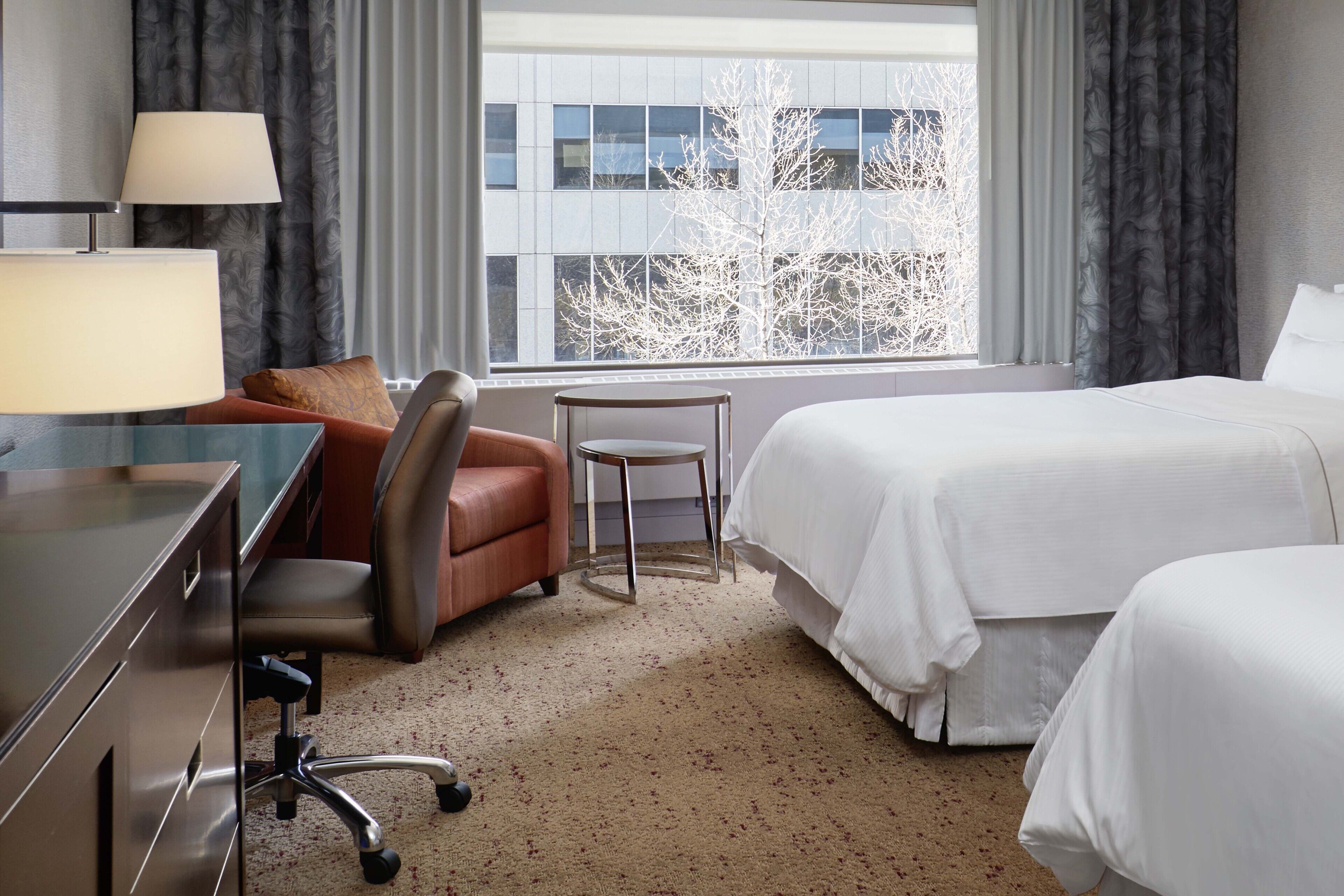 A hotel room with a desk, two chairs, a bed, and a window with a view of buildings and trees.