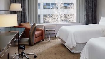 A hotel room with a desk, two chairs, a bed, and a window with a view of buildings and trees.