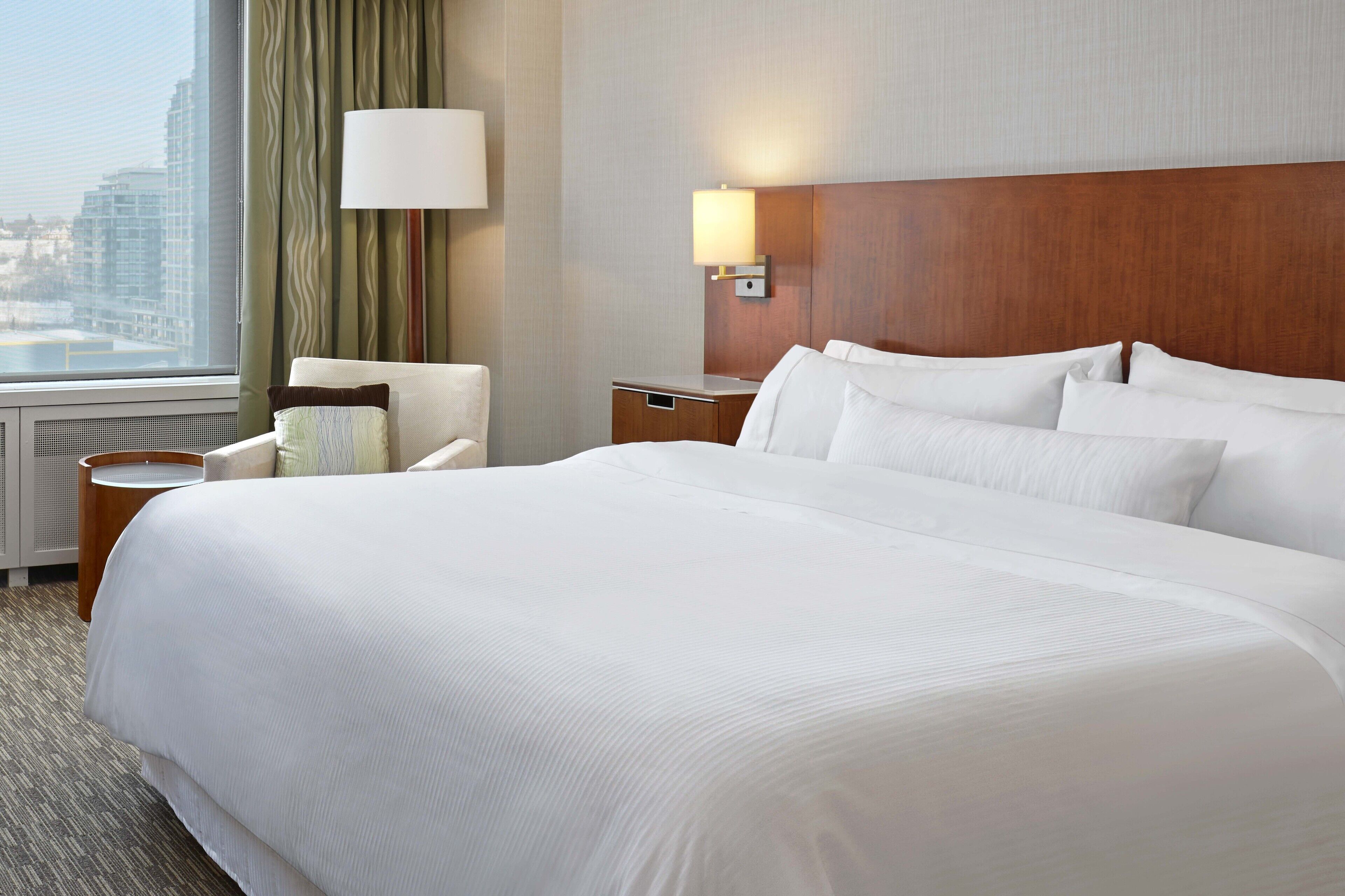 Habitación de hotel con una cama grande, una mesita de noche, una lámpara, una silla y vistas a la ciudad.