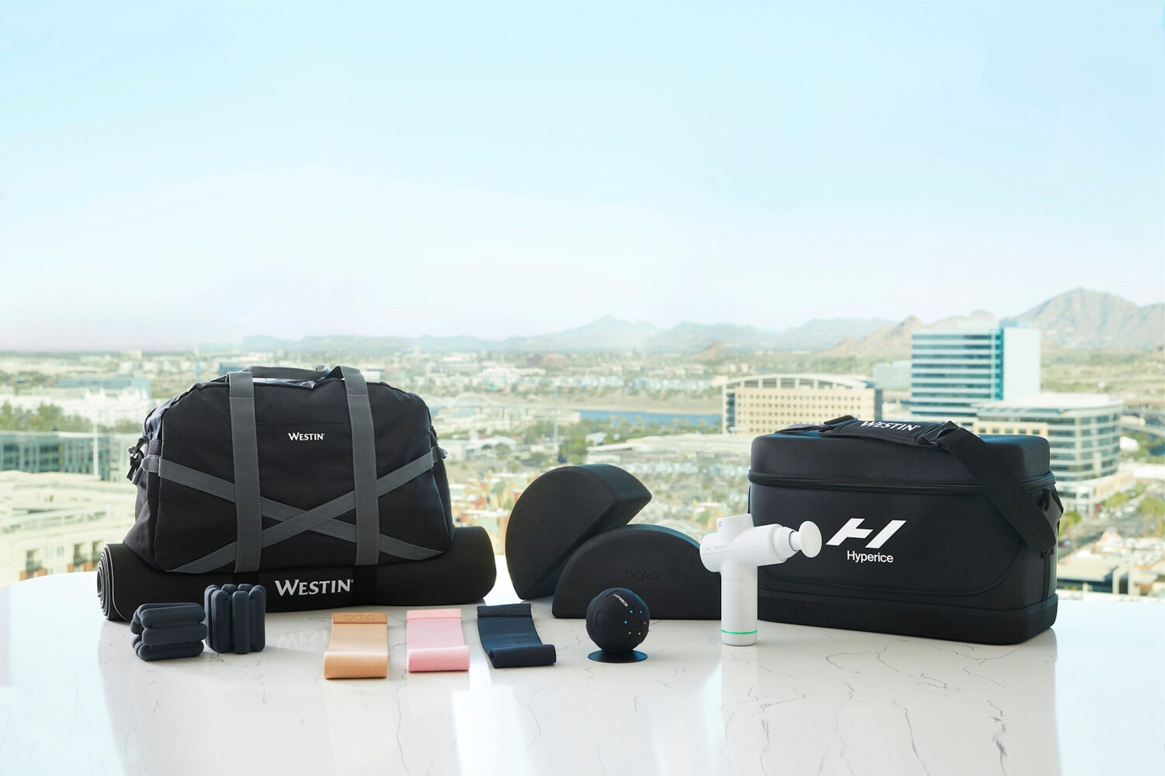 A Westin Hyperice massage device, a Westin branded massage bag, and a massage ball on a table with a city view in the background.