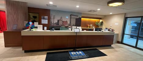 A hotel reception desk with a cityscape mural, a digital screen, and a black mat with "RH" on it.
