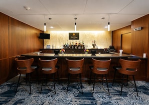 A bar area with a marble countertop, multiple bar stools, a television mounted on the wall, and a selection of bottles and glasses.