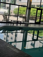 Una piscina interior con paredes de vidrio transparente, suelo de baldosas y vistas a edificios y vegetación.