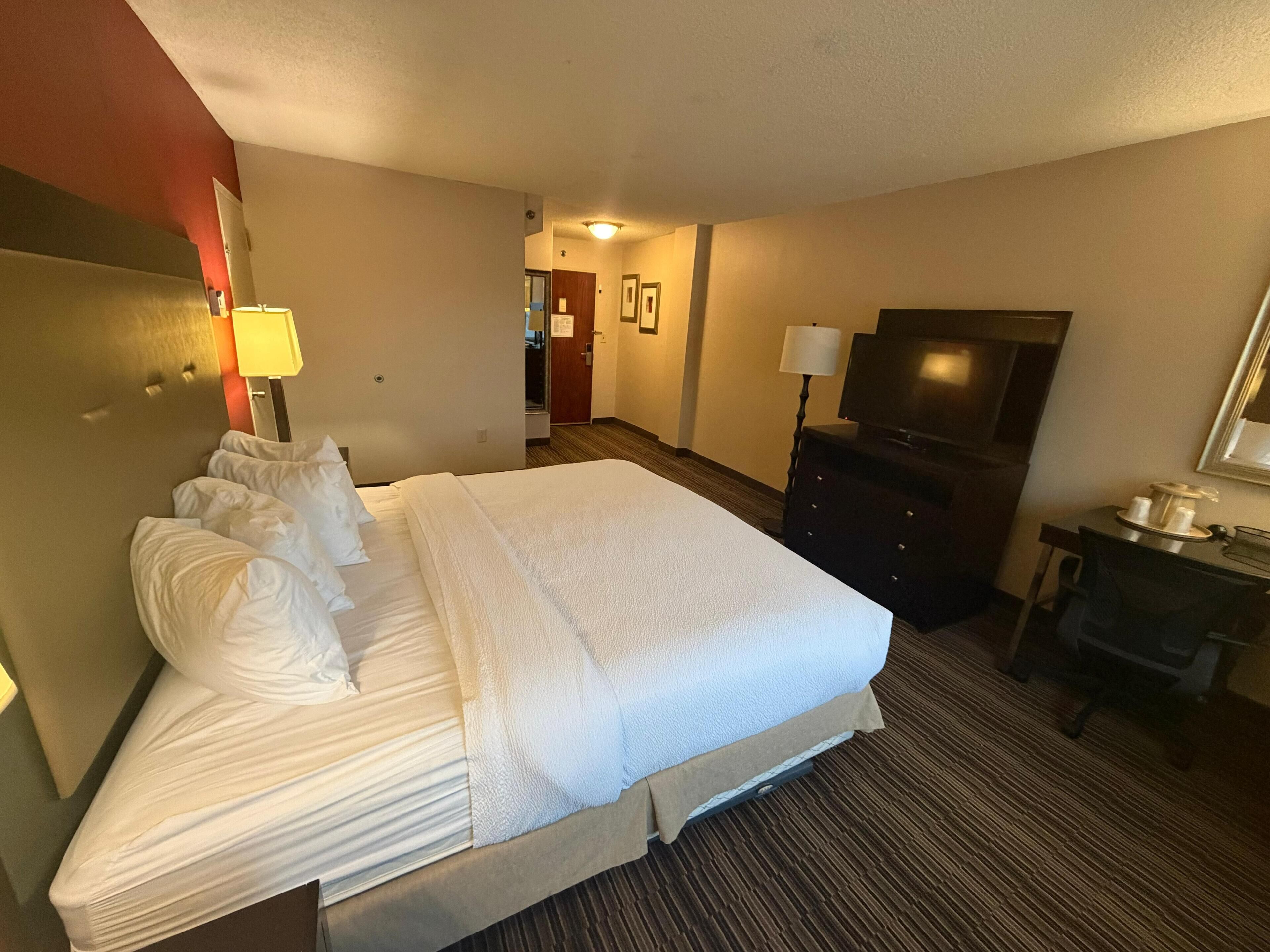 A hotel room with a large bed, a dresser, a TV, and a desk with a lamp.