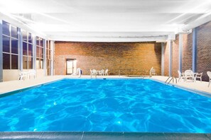 An indoor swimming pool with clear blue water, surrounded by white chairs and tables, and a brick wall in the background.