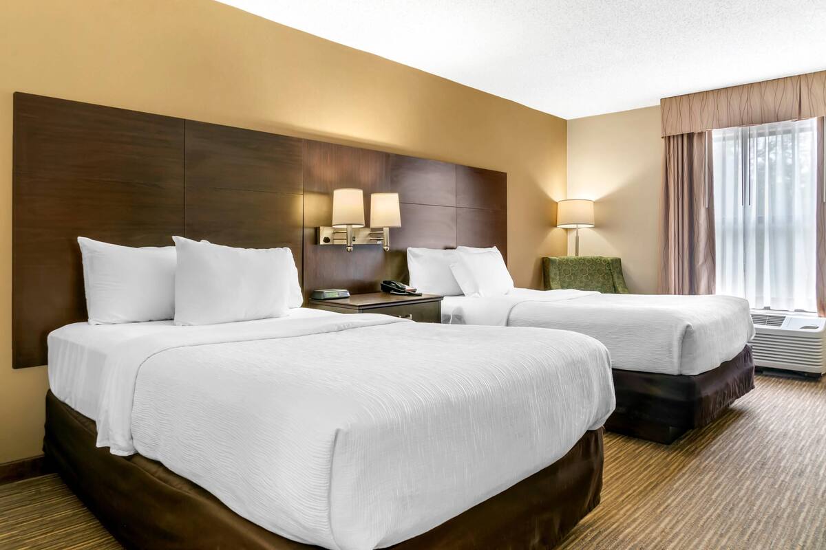 A hotel room with two beds, a wooden headboard, bedside lamps, a nightstand, and a window with curtains.