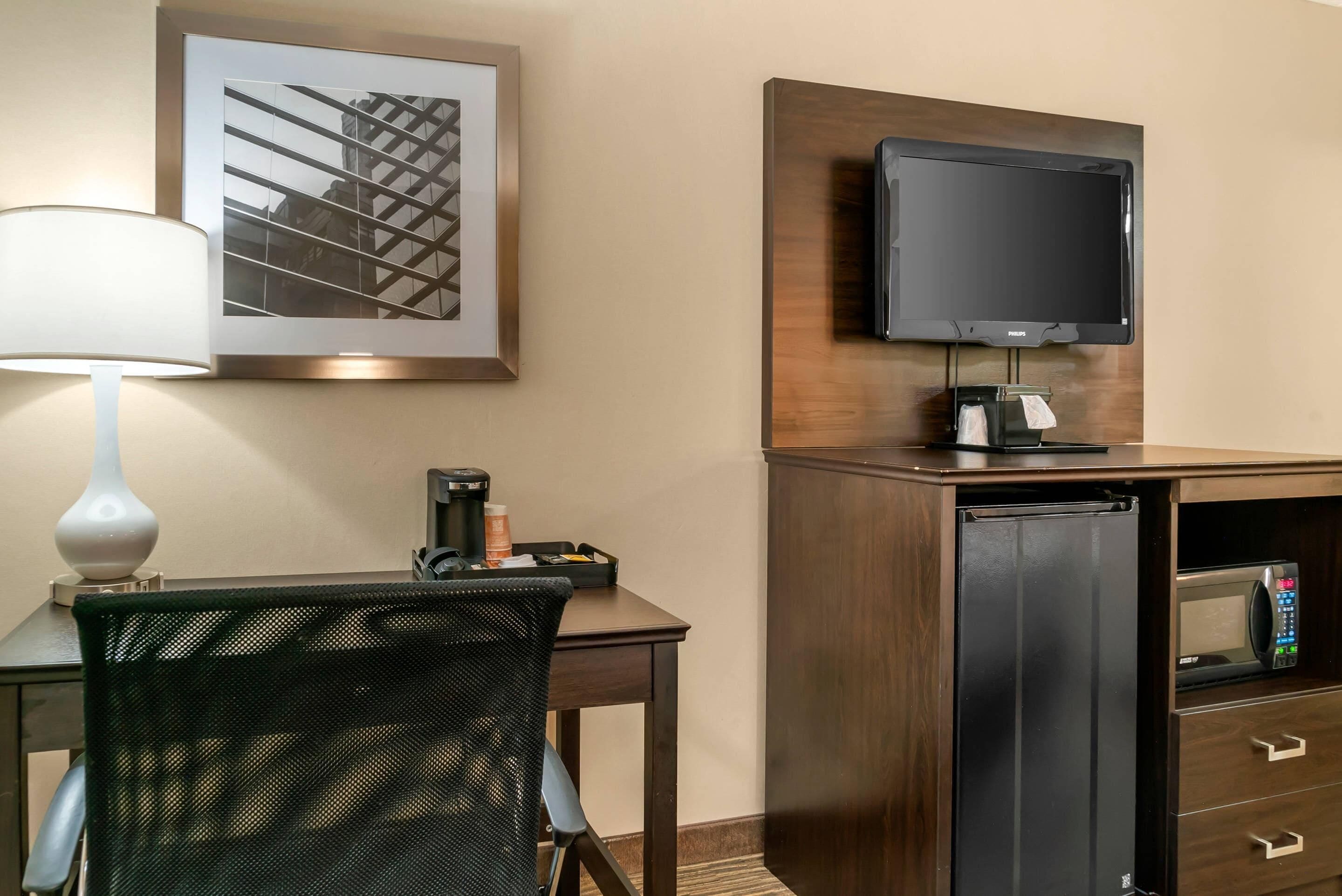 A hotel room with a desk, chair, lamp, flat-screen TV, minibar, and a framed picture on the wall.
