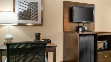 A hotel room with a desk, chair, lamp, flat-screen TV, minibar, and a framed picture on the wall.