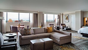 Habitación de hotel con un amplio sofá modular, una mesa de centro redonda y una zona de comedor con sillas y una mesa. Hay una cama con un ventanal y vistas a la ciudad.
