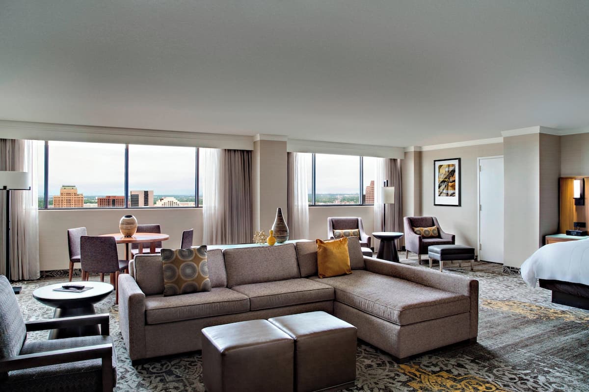 Une chambre d’hôtel dotée d’un grand canapé d’angle, d’une table basse ronde et d’un coin repas avec des chaises et une table. On y trouve un lit avec une grande fenêtre donnant sur le paysage urbain.