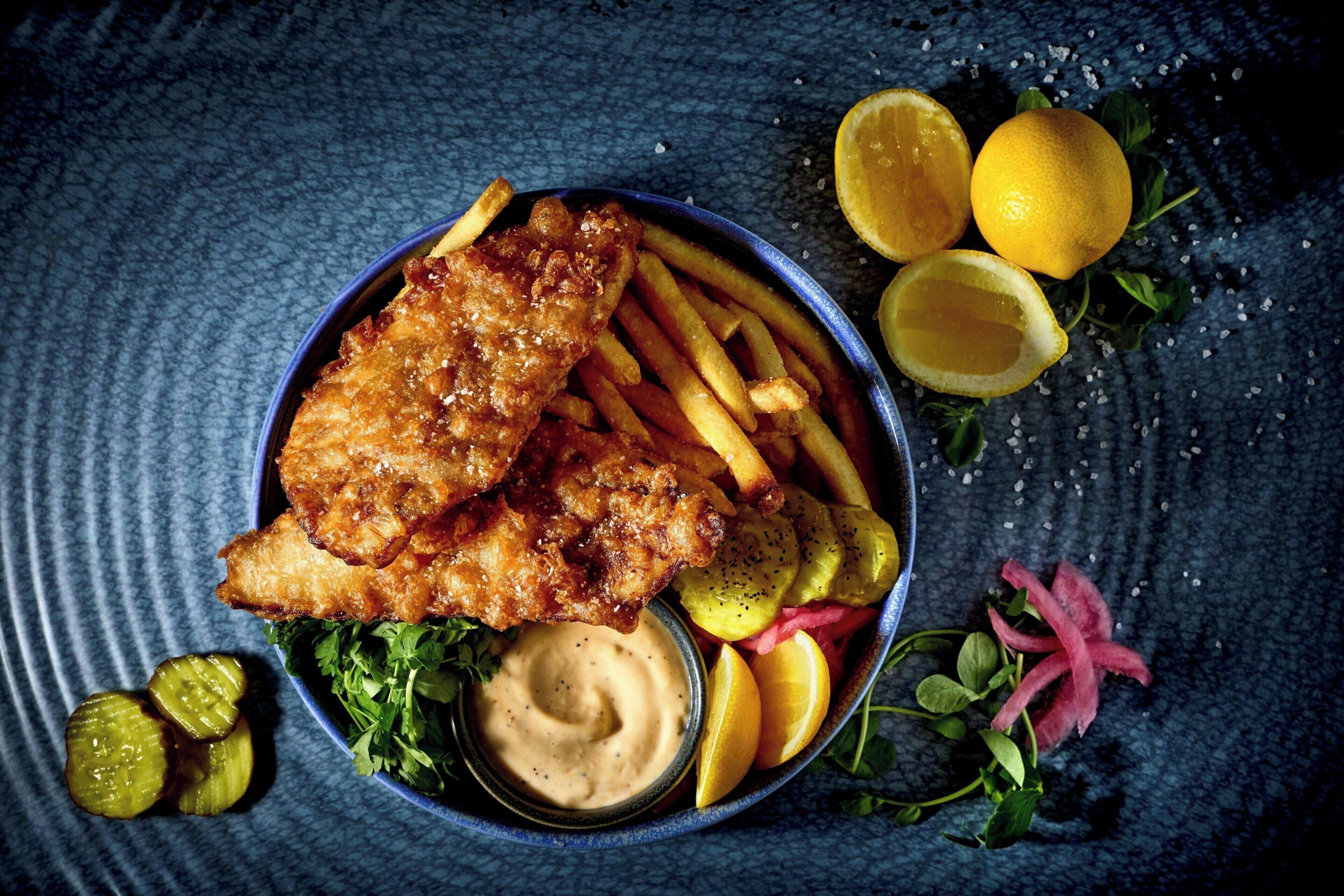A bowl of fish and chips with fries, lemon slices, pickles, and a dipping sauce.