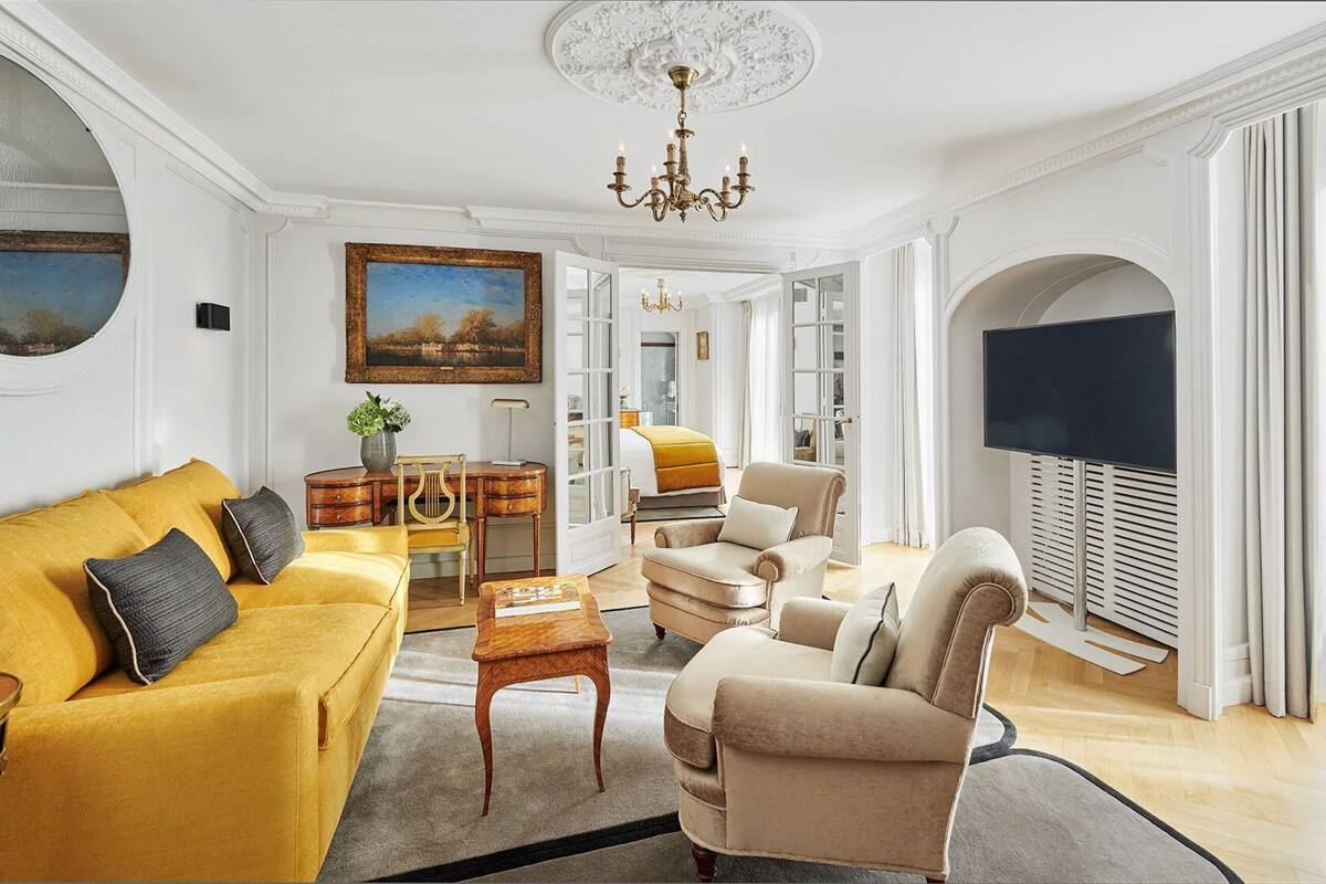 Un salón espacioso con un sofá amarillo, un sillón color beige y una mesa de centro de madera. Un candelabro cuelga del techo, y hay un cuadro en la pared.