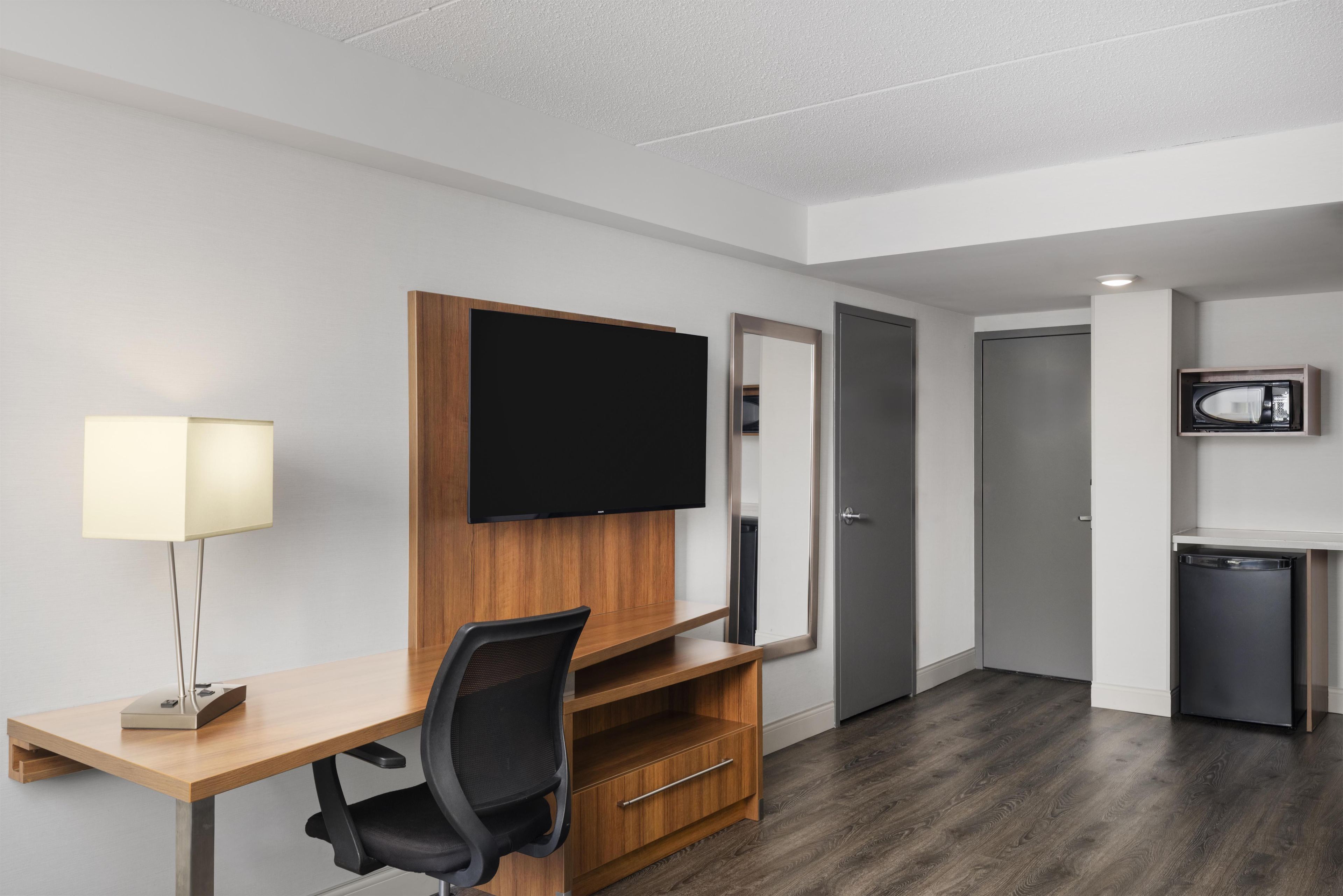 A hotel room with a wooden desk, a chair, a television mounted on the wall, a door, a microwave, and a small refrigerator.