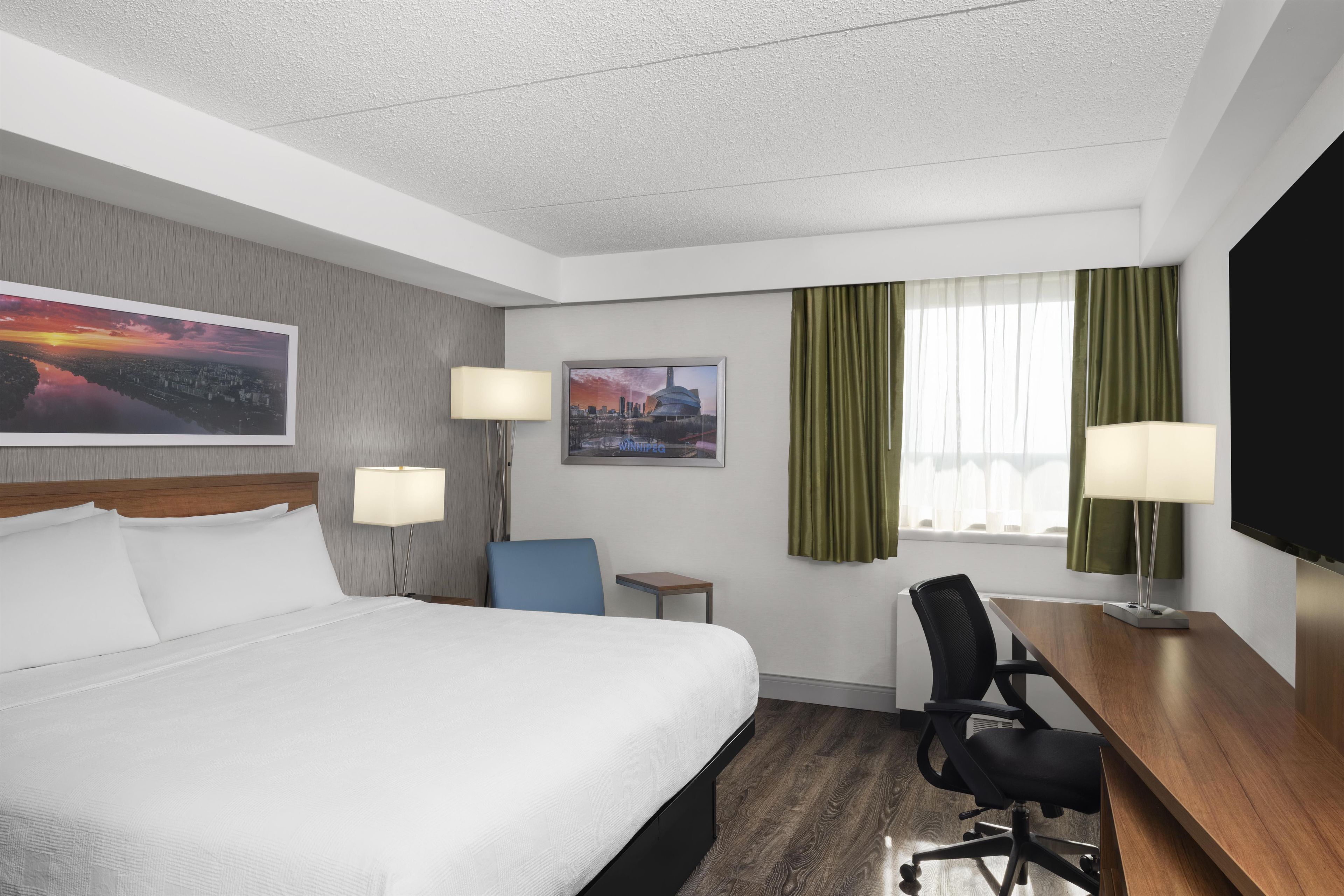 A hotel room with a large bed, two bedside tables, a desk with a chair, a TV, and two windows with curtains.