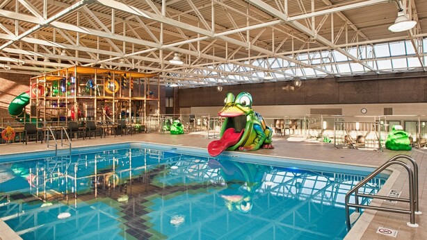 Una piscina interior con un tobogán de rana, rodeada por un techo alto con vigas metálicas y una zona de juegos con equipo colorido.