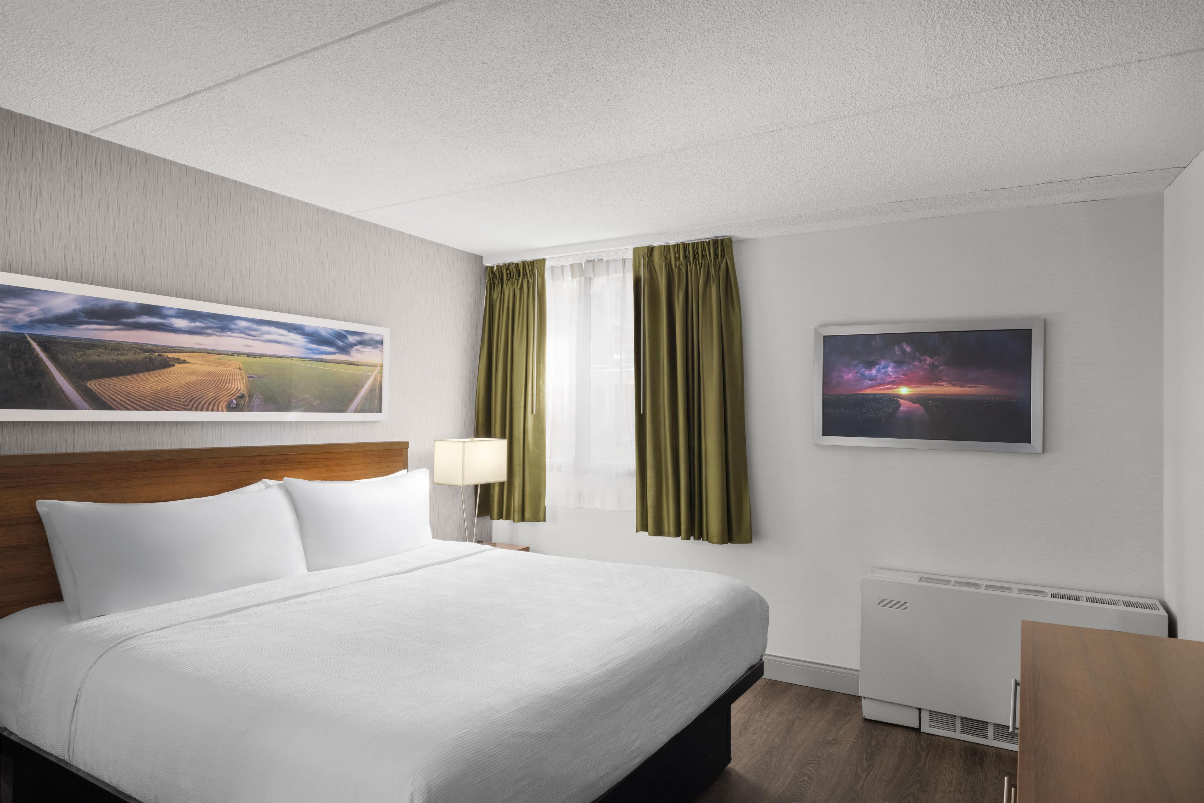 A hotel room with a bed, a nightstand, a lamp, a window with curtains, and two framed pictures on the wall.