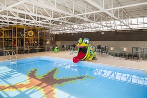 Una piscina interior con tobogán, rodeada por una zona de juegos con estructuras para escalar y un área de descanso con mesas y sillas.