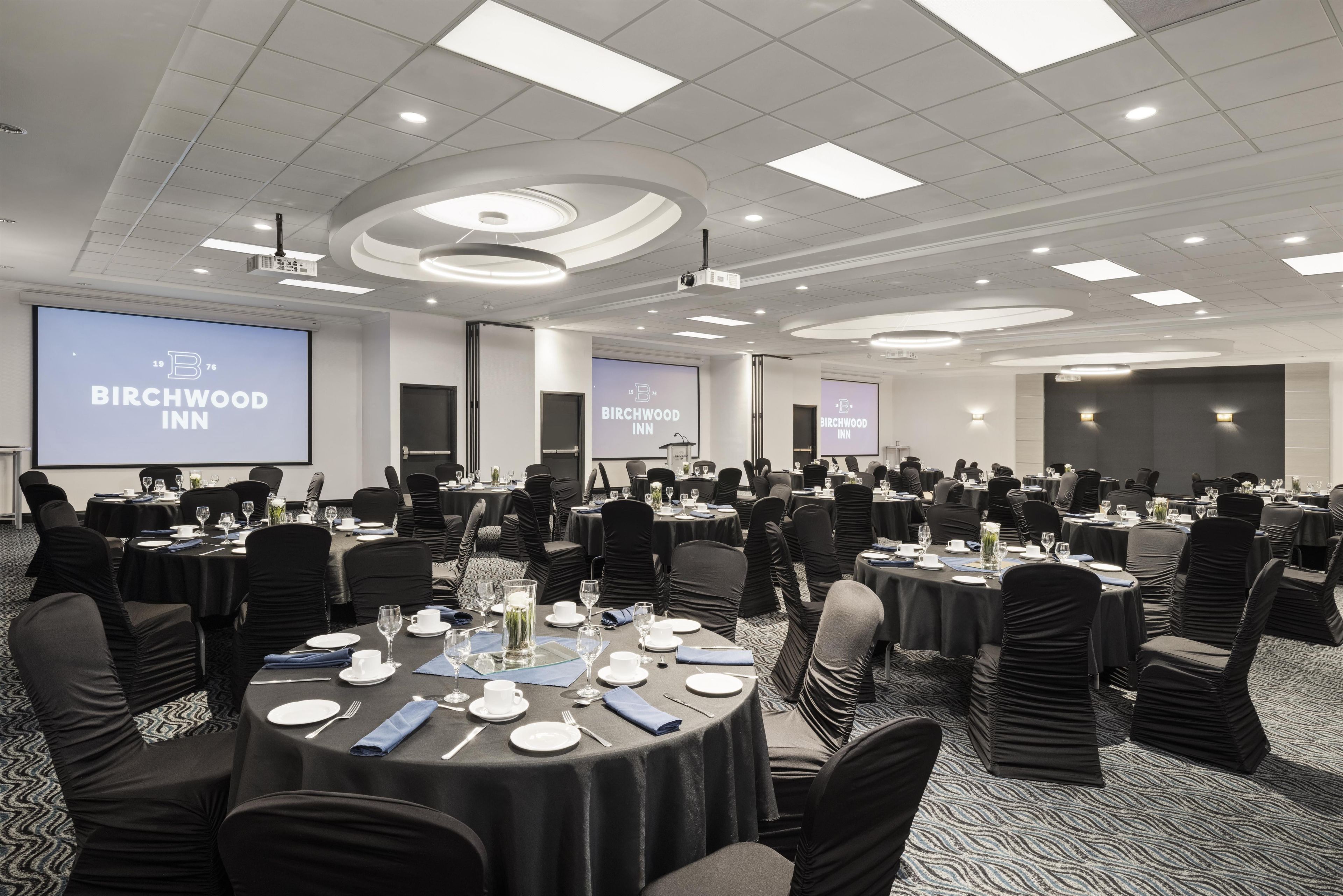 Sala de conferências com várias mesas preparadas para uma reunião, equipada com projetores e telas exibindo o nome do hotel.