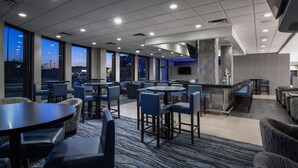 Un moderno vestíbulo de hotel con mesas redondas y sillas azules, amplias ventanas con vistas a la ciudad y una zona de bar.