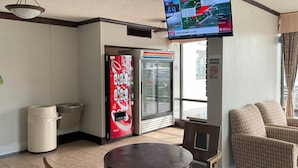 A room with a television displaying a weather forecast, a Coca-Cola vending machine, a trash bin, a round table with two chairs, and a sofa.