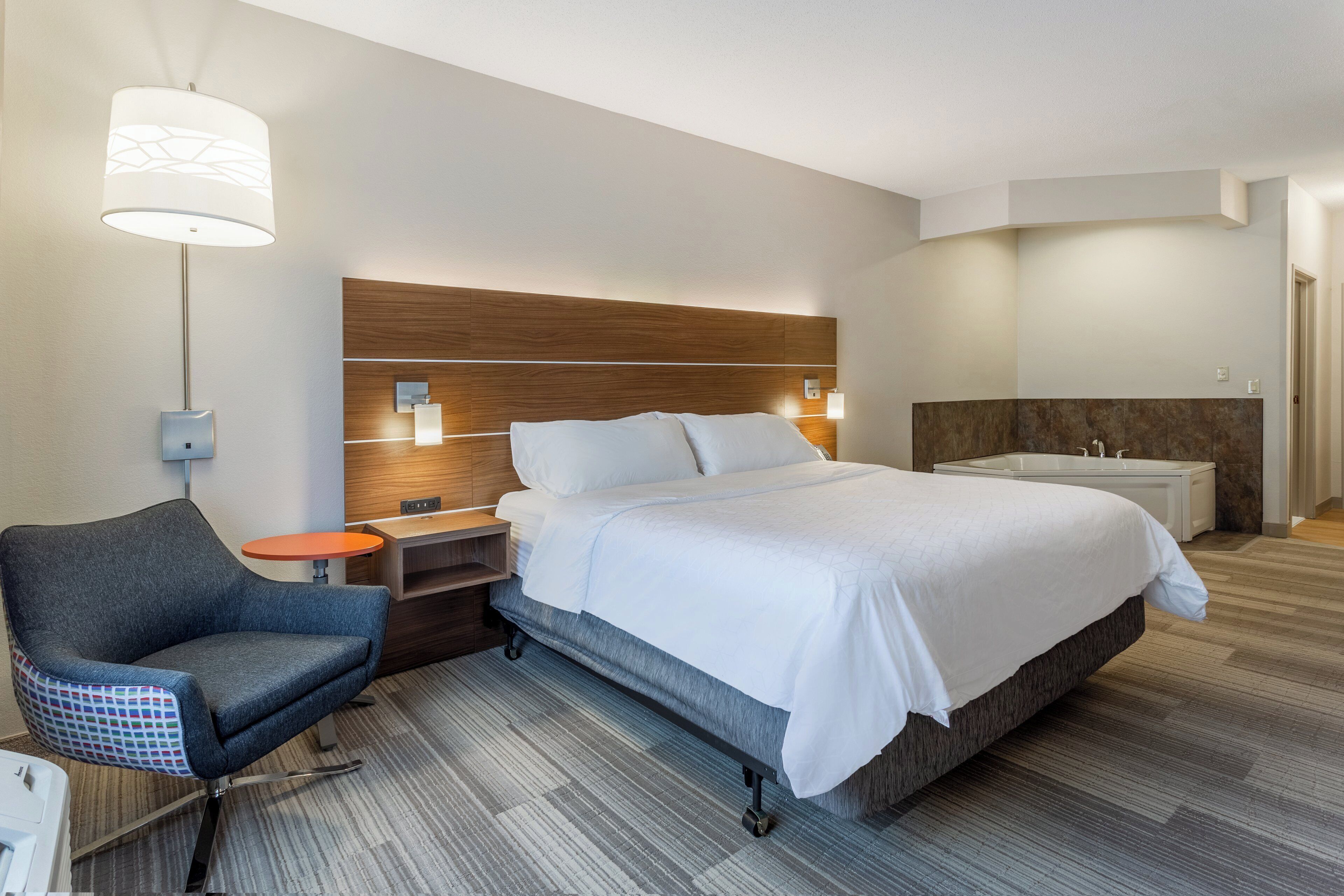 A hotel room with a bed, a chair, a nightstand, a lamp, and a bathroom.
