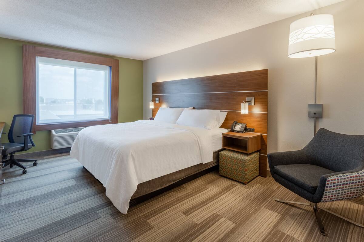 A hotel room with a bed, a desk, a chair, a lamp, and a window.