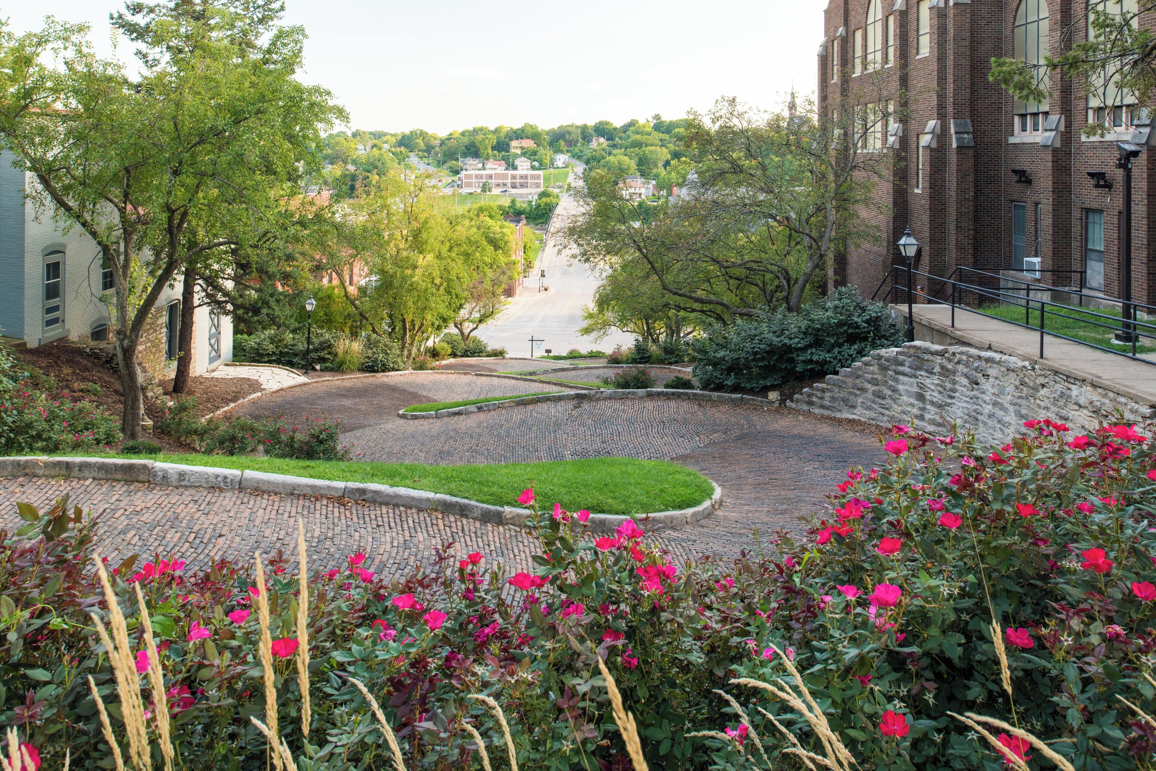 A paved courtyard with a curved walkway, surrounded by trees and flowering plants.