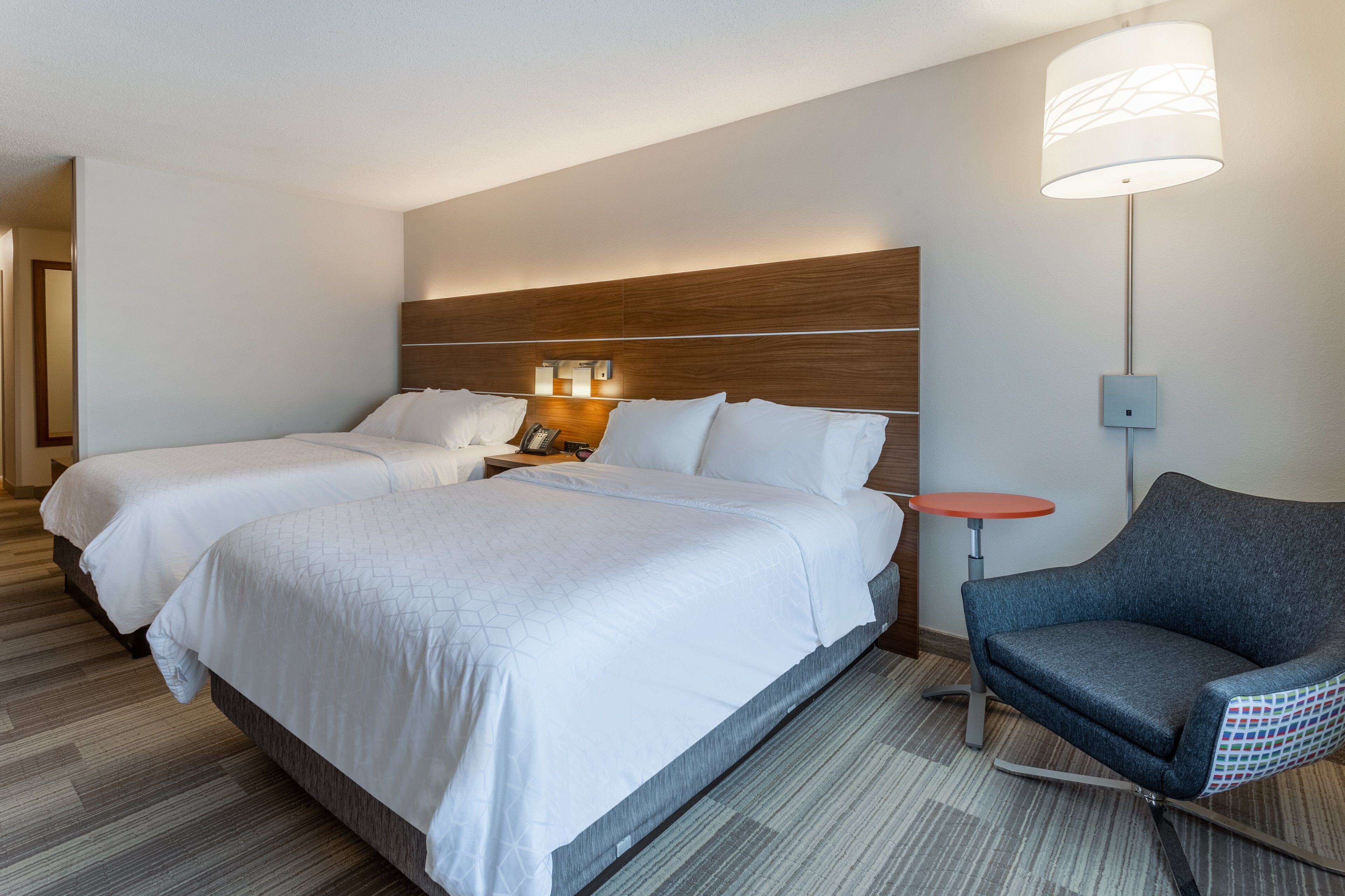 A hotel room with two beds, a nightstand, a lamp, and a chair.