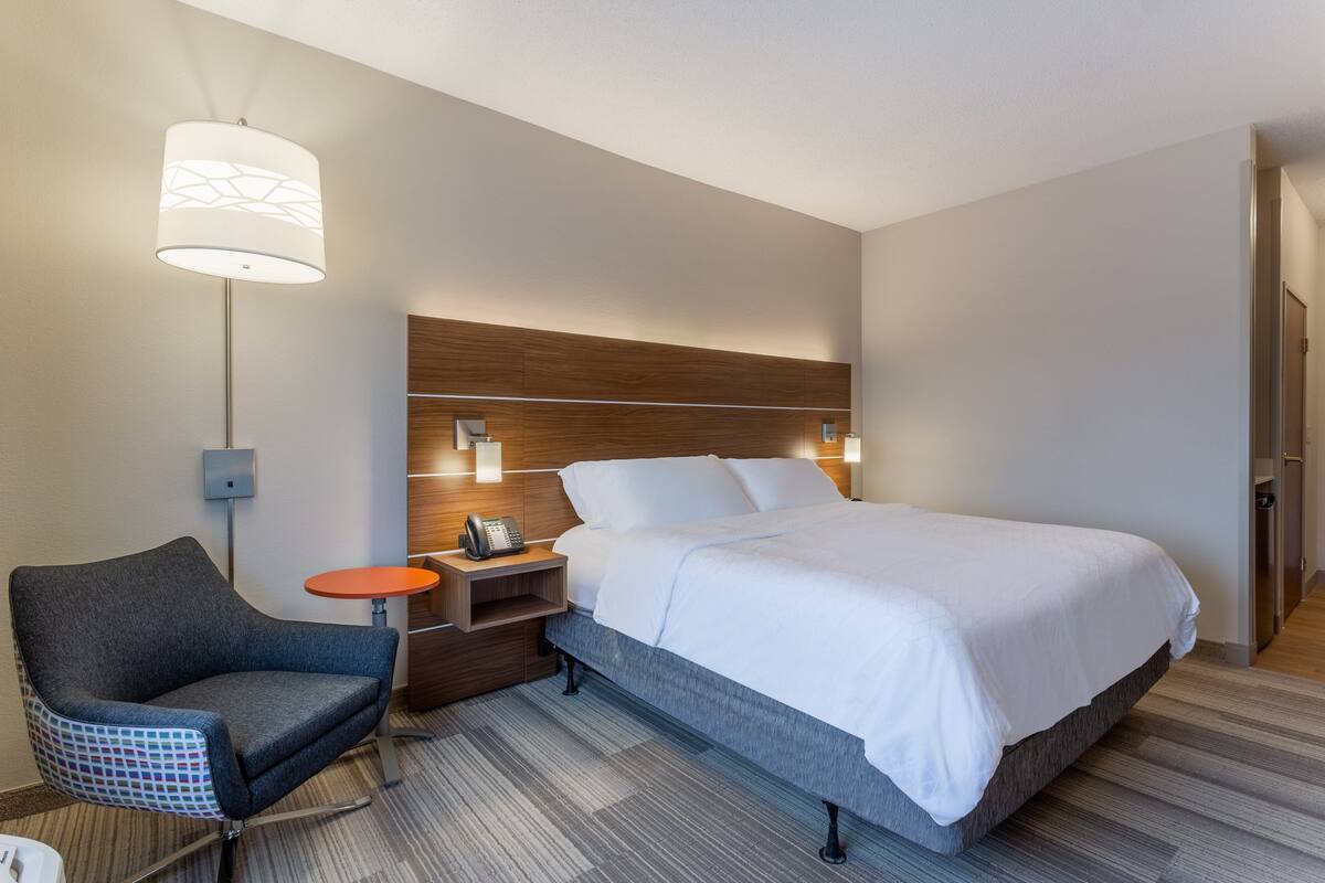 A hotel room with a large bed, a chair, a nightstand with a phone, and a wall-mounted lamp.