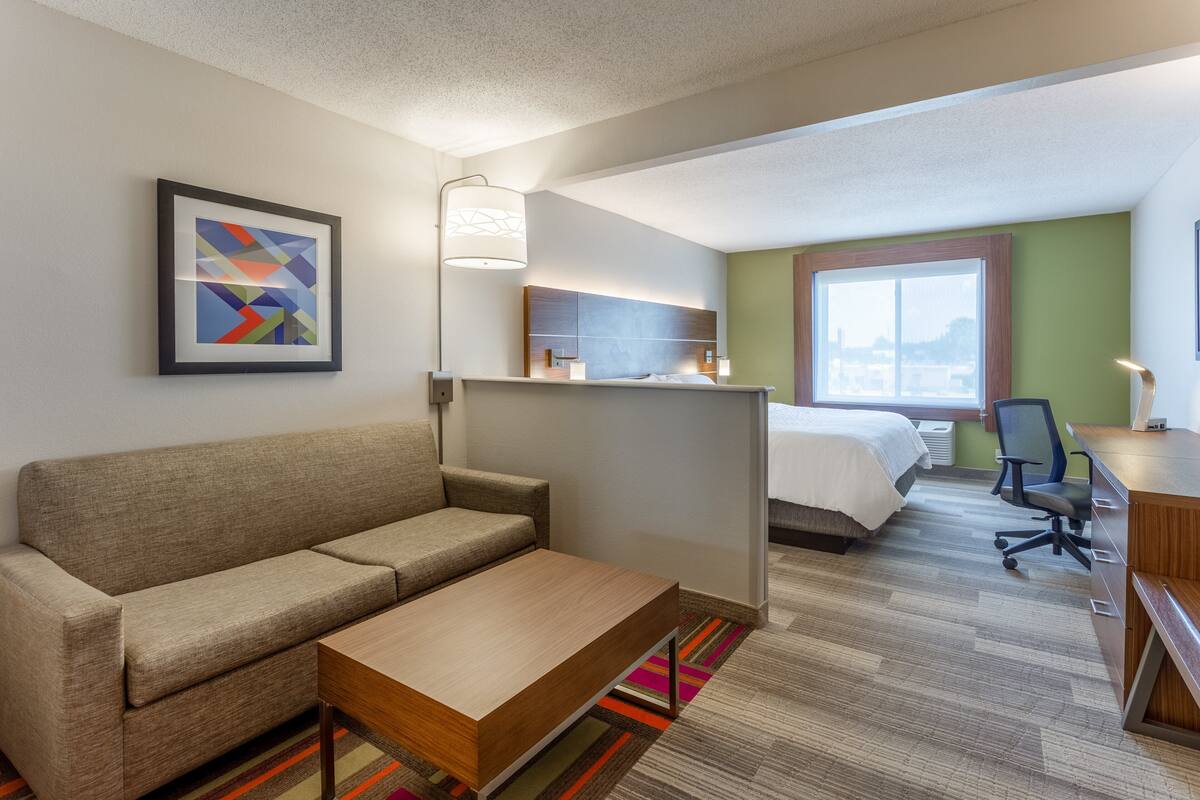 A hotel room with a sofa, a wooden coffee table, a desk with a chair, a bed, a window, and a framed abstract painting on the wall.