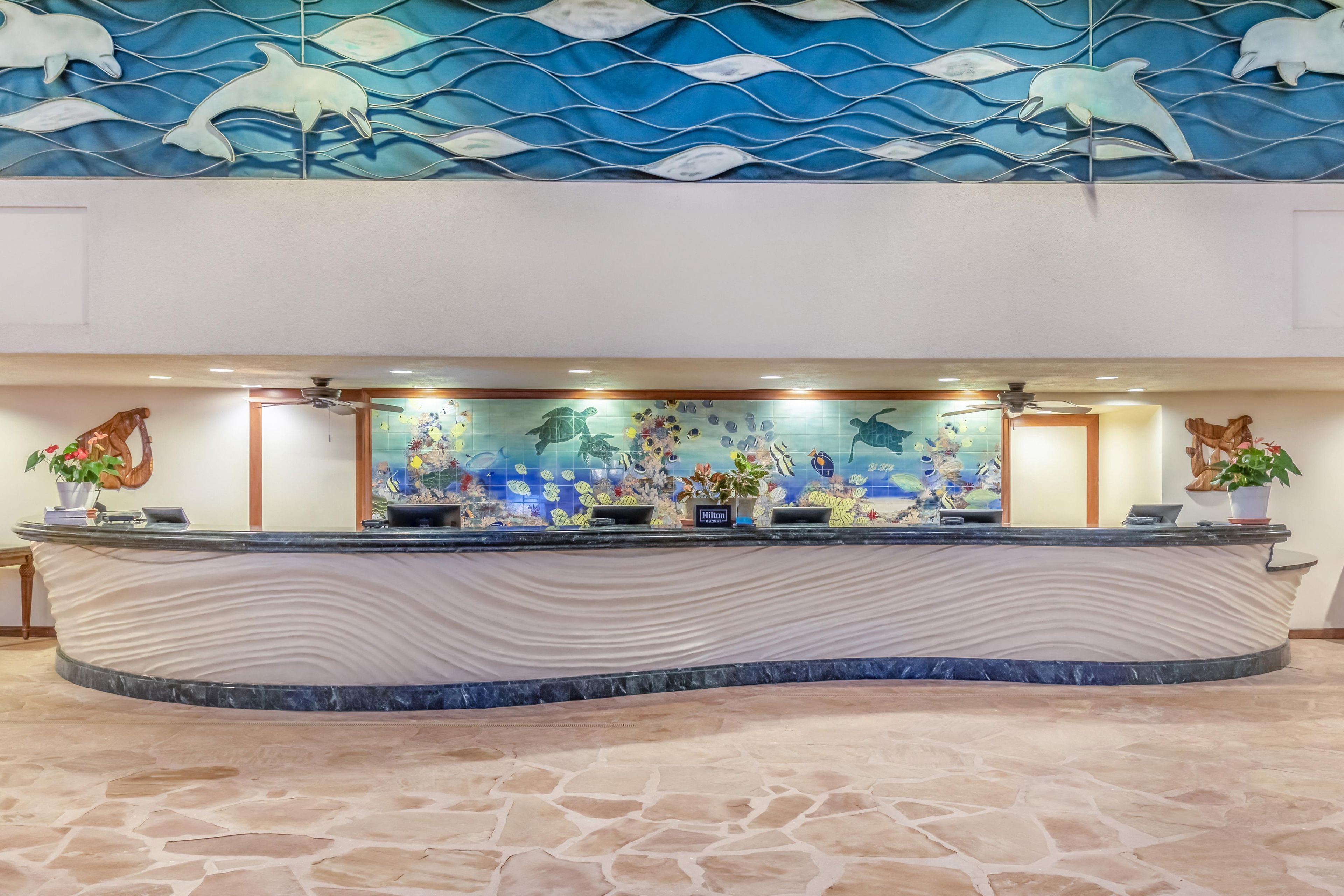 A hotel reception desk with a curved marble top, a large aquarium with various fish and a turtle, and a mural of dolphins on the wall above.