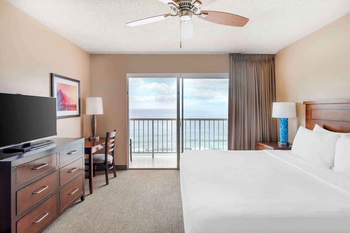 A hotel room with a bed, a TV, a desk with a chair, a ceiling fan, and a view of the ocean through a large window.