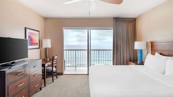 Habitación de hotel con cama, televisor, escritorio con silla, ventilador de techo y vista al océano a través de un gran ventanal.