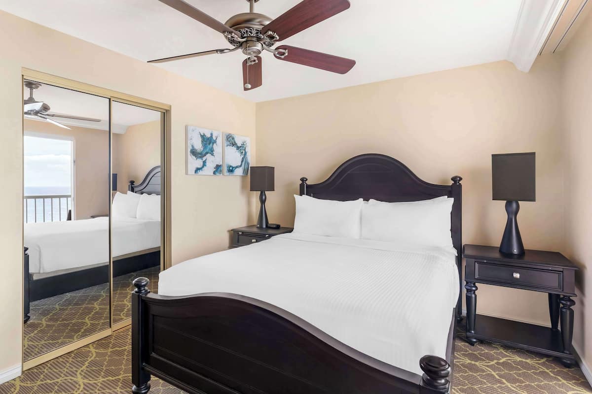 A hotel room with a large bed, a ceiling fan, a nightstand with a lamp, a mirror, and artwork on the wall.