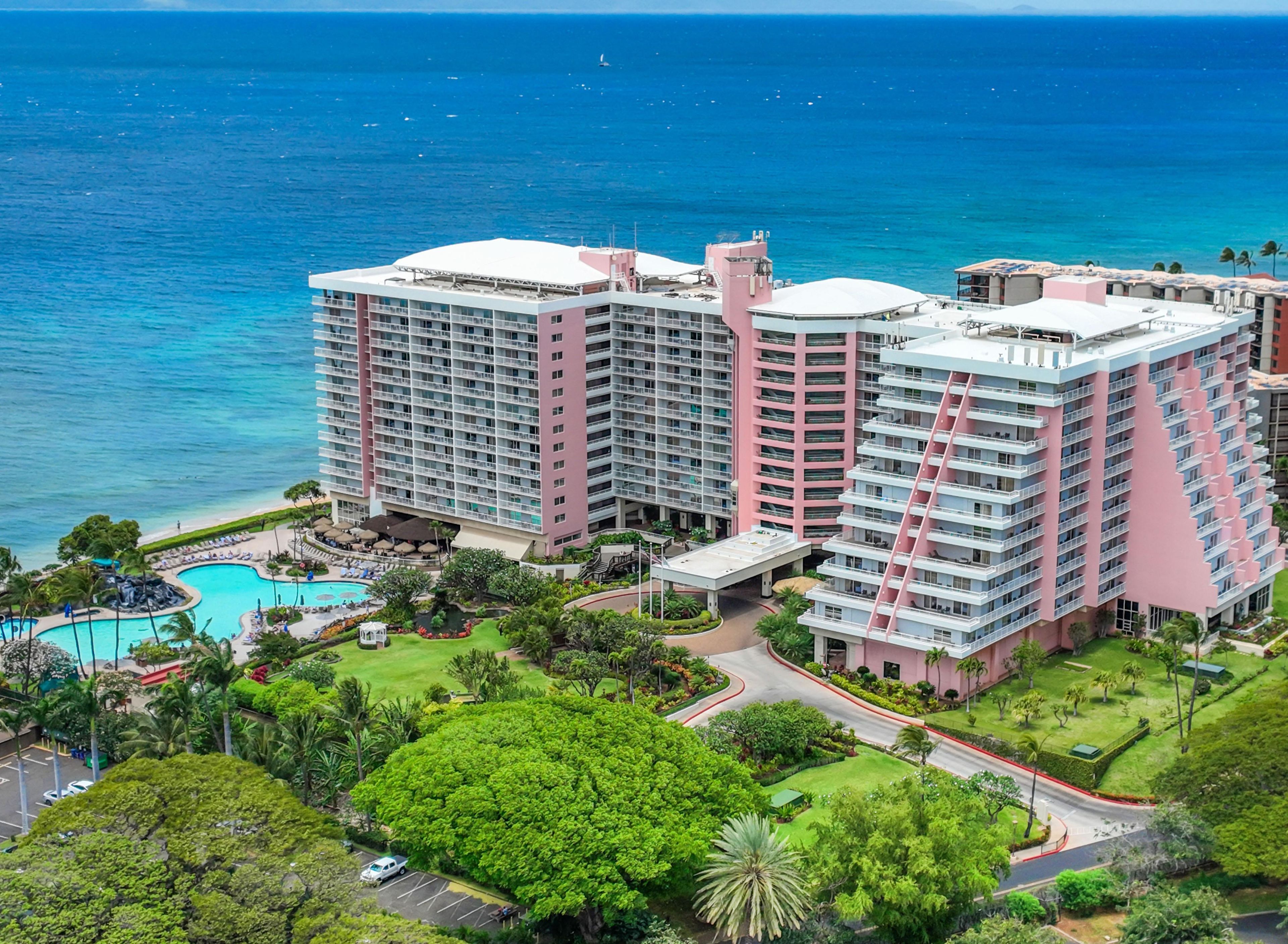 A multi-story hotel with a pool, surrounded by lush greenery and a view of the ocean.