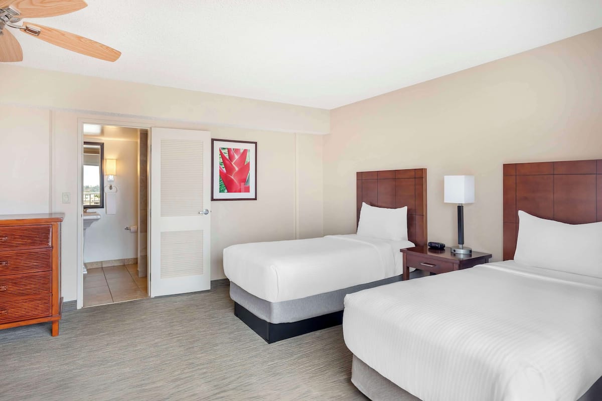A hotel room with two beds, a wooden dresser, a ceiling fan, and a bathroom visible through an open door.