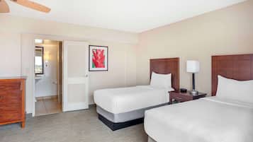 Quarto de hotel com duas camas, cômoda de madeira, ventoinha no teto e banheiro visível através de uma porta aberta.